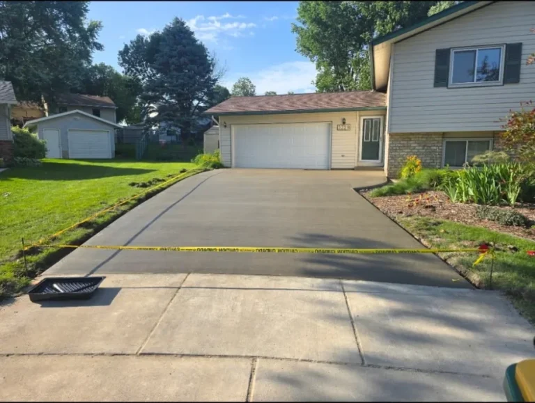 Fresh gray concrete driveway leading up to the two-car garage of a tan split-level home, yellow caution tape strung across the bottom where it meets the older sidewalk panels, Byron, MN