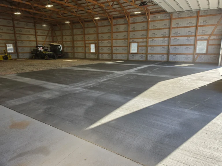 Inside a large pole building with exposed wood trusses and house-wrapped walls: a fresh gray concrete floor with a saw-cut joint grid covers the front of the building, sunlight falls across it from the open door, and the back of the building is still a compacted gravel floor where two utility vehicles are parked, near Medford, MN